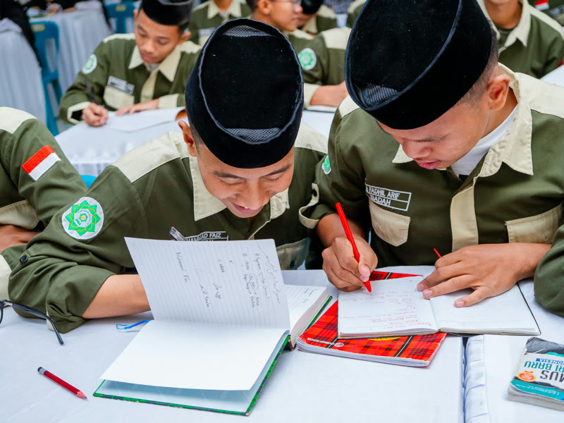 Dua santri berseragam hijau dan berpeci hitam menulis catatan di buku tulis, berdampingan di meja belajar