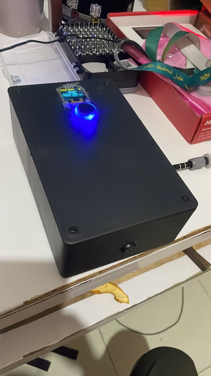 Unit absensi di meja kerja: kotak hitam dengan layar OLED bertuliskan Siap dan sensor sidik jari bulat yang menyala biru