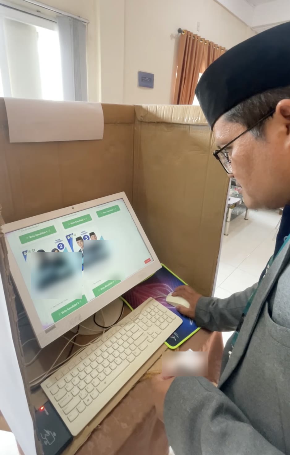 Ustadz memilih di bilik suara berdinding kardus, layar menampilkan kartu kandidat nomor 2 dan 3 dengan tombol Vote Kandidat