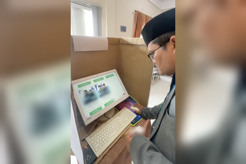 Seorang ustadz memilih di bilik suara berdinding kardus, layar komputer menampilkan kartu pasangan kandidat OSIA